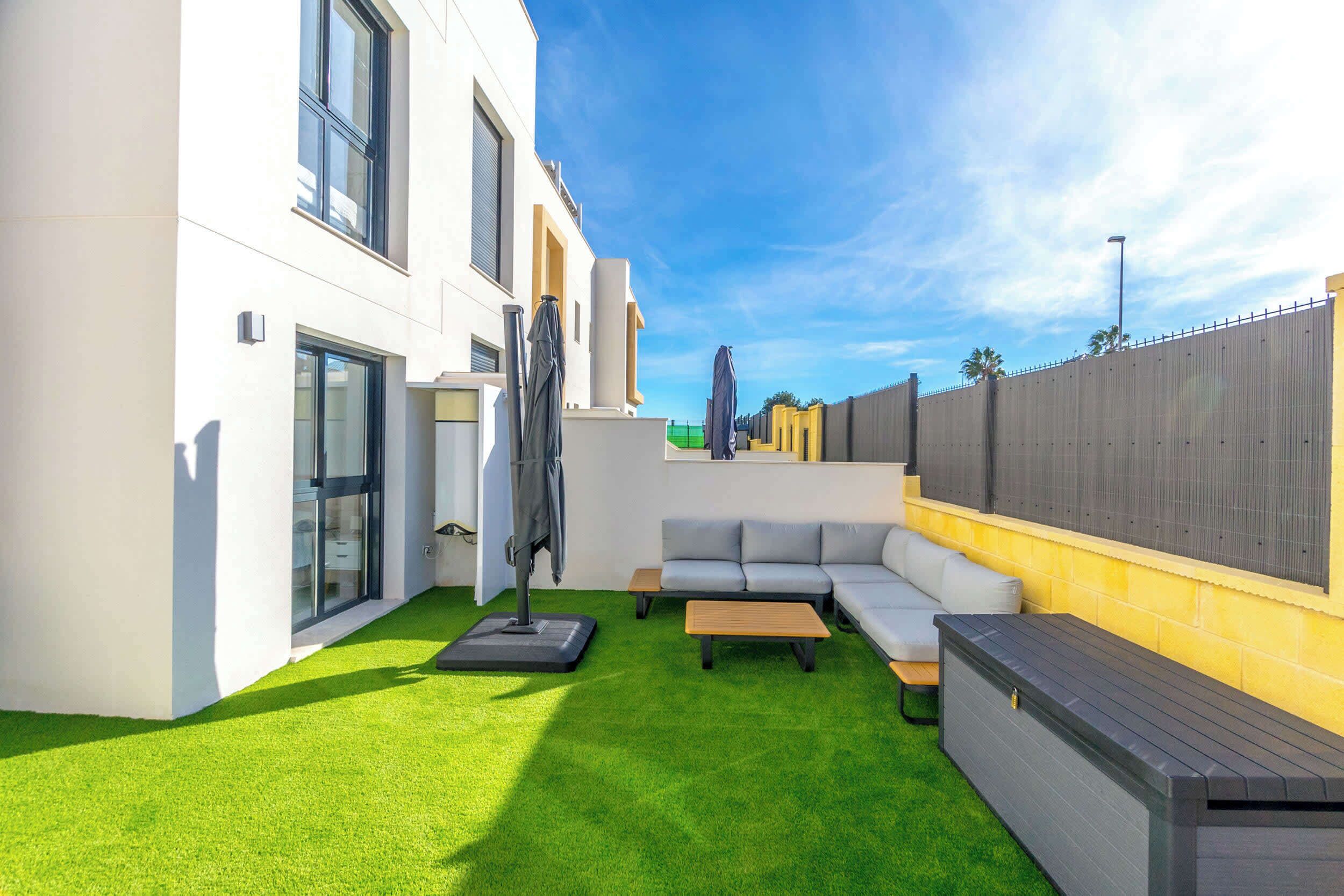 A modern outdoor patio with a green lawn, a grey sofa, and a wooden table.
