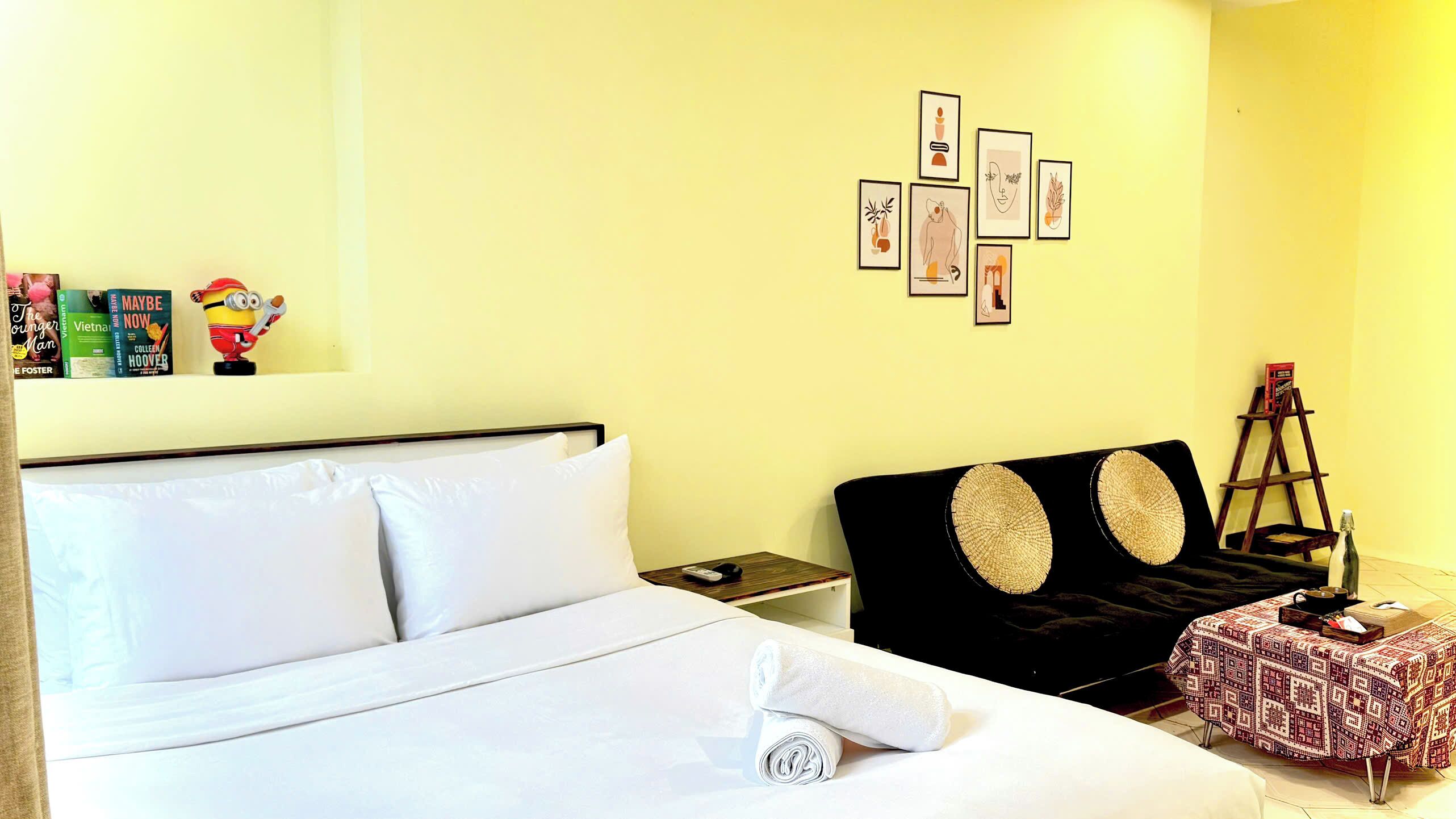 A hotel room with a bed, a nightstand, a black headboard, a small table with a bottle, and a ladder against the wall.