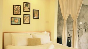 Una cama bien tendida con sábanas blancas y cabecera de madera, una pared con obras de arte enmarcadas y una ventana con cortinas traslúcidas.