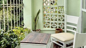 Un balcón pequeño y cerrado con una silla blanca y una mesita, rodeado de plantas en macetas y un separador decorativo.