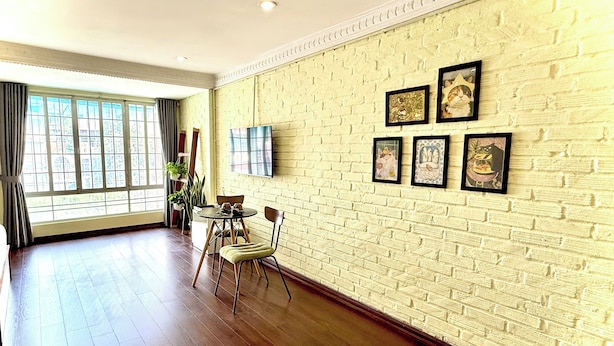 Una habitación amplia con pared de ladrillo, suelo de madera, zona de comedor con sillas y una mesa, y un gran ventanal con cortinas.
