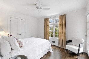 A bedroom with a bed, a chair, a window with curtains, a ceiling fan, and a door.
