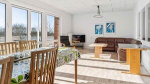 A spacious living area with a dining table, chairs, a sofa, and a TV. Large windows provide ample natural light.