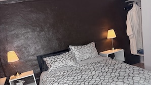A modern bedroom with a dark wall, a patterned bedspread, two bedside lamps, and a white robe hanging on a rack.