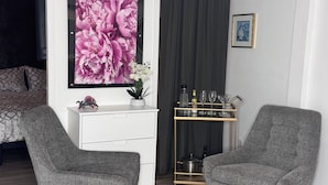 A modern living room with two grey armchairs, a white console table with a decorative item, a large floral artwork, a gold-framed picture on the wall, and a small shelf with bottles and glasses.
