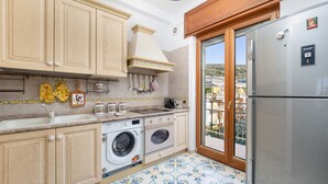 A kitchen with wooden cabinets, a washing machine, a refrigerator, and a view of a hillside through a glass door. - Casa Cafarelli - Bright apt with Stunning view (Sorrento)