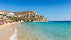 Ein Strand mit klarem türkisblauem Wasser, einem sandigen Ufer und Gebäuden, die sich einen Hügel hinaufziehen.