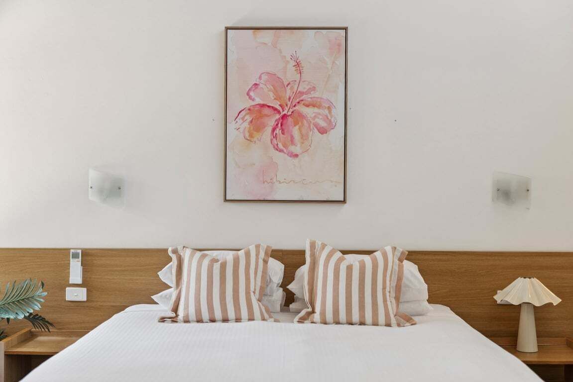 Habitación de hotel con cabecera de madera, cama con almohadas a rayas, un cuadro colgado en la pared de una flor de hibisco y lámparas de noche.