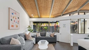 A modern living room with a grey sofa, a white coffee table, and a large window with a view of greenery outside. - Hibiscus Villa - Pet Friendly, Spacious beachfront studio, private courtyard (Coffs Harbour)