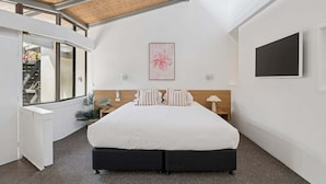 A modern bedroom with a large bed, a wooden headboard, a flat-screen TV, and a window overlooking a balcony. - Hibiscus Villa - Pet Friendly, Spacious beachfront studio, private courtyard (Coffs Harbour)