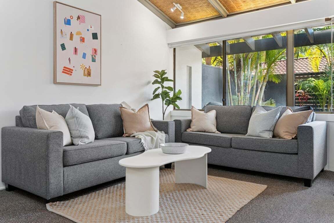 Een moderne woonkamer met twee grijze banken, een witte salontafel en een groot raam met uitzicht op groen.