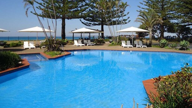 A swimming pool with lounge chairs and umbrellas, surrounded by tall trees and a view of the ocean. - Hibiscus Villa - Pet Friendly, Spacious beachfront studio, private courtyard (Coffs Harbour)