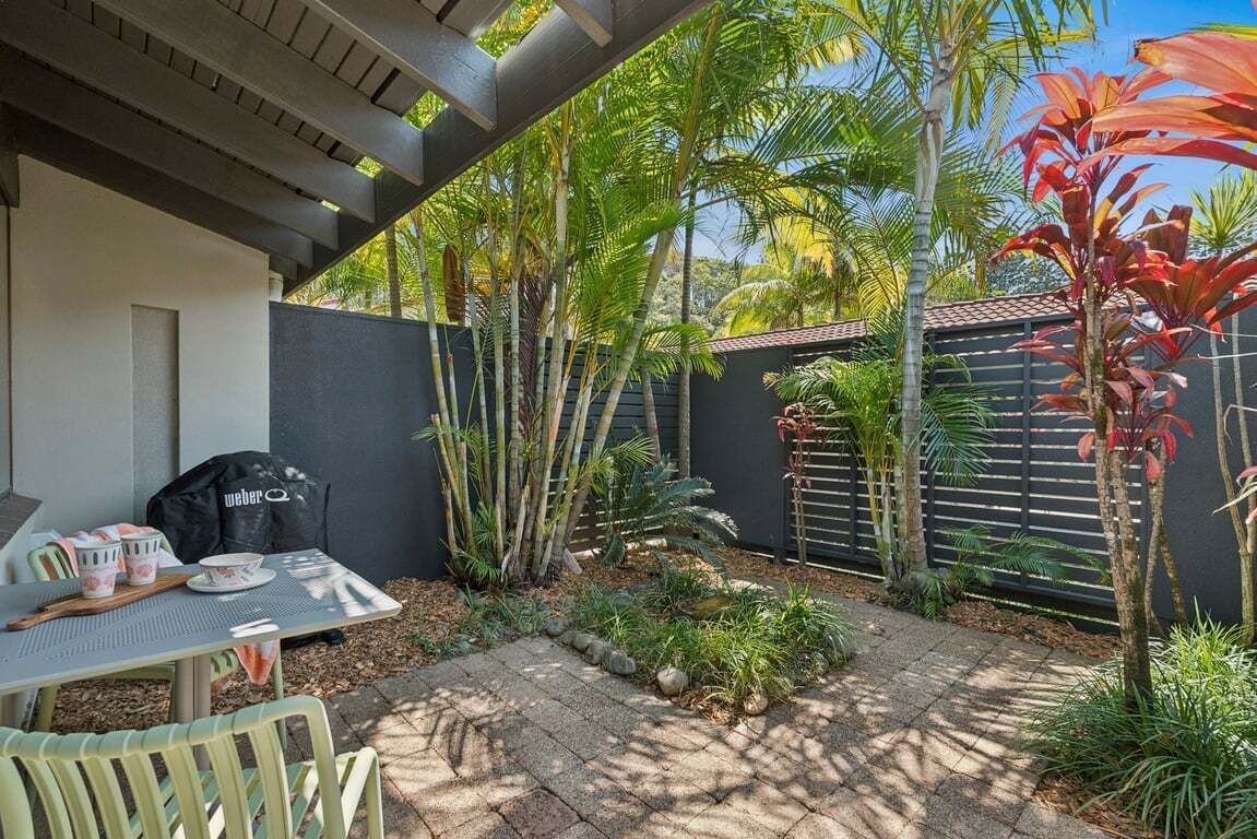 A patio area with a table and chairs, surrounded by tropical plants and a fence.