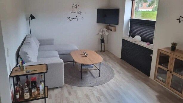 Ein modernes Wohnzimmer mit einem grauen Sofa, einem Holztisch, einem an der Wand montierten Fernseher und einer kleinen Kommode.