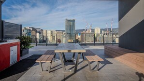 A rooftop terrace with a table and benches, offering a city view with buildings and cranes in the background.
