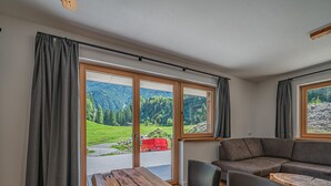 A spacious living area with a wooden table, two chairs, a sofa, and a coffee table. Large windows offer a view of a green landscape and a red truck outside. - Two Alpine flats with charm (Wald im Pinzgau)
