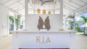 A reception area with a white counter, the name "RIA" and "ULUWATU" displayed, and a decorative winged sculpture above. - RIA ULUWATU Tropical Chic Hotel (Pecatu)