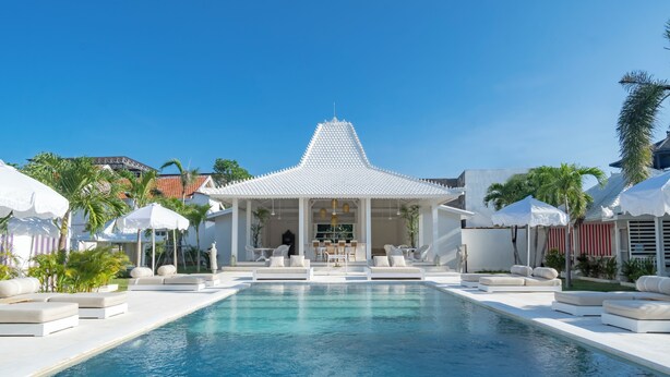 A swimming pool with white lounge chairs and umbrellas, a covered seating area with a tiled roof, and palm trees. - RIA ULUWATU Tropical Chic Hotel (Pecatu)