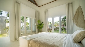 A large bed with a beige blanket, a bedside table, and a view of a garden through large windows. - RIA ULUWATU Tropical Chic Hotel (Pecatu)