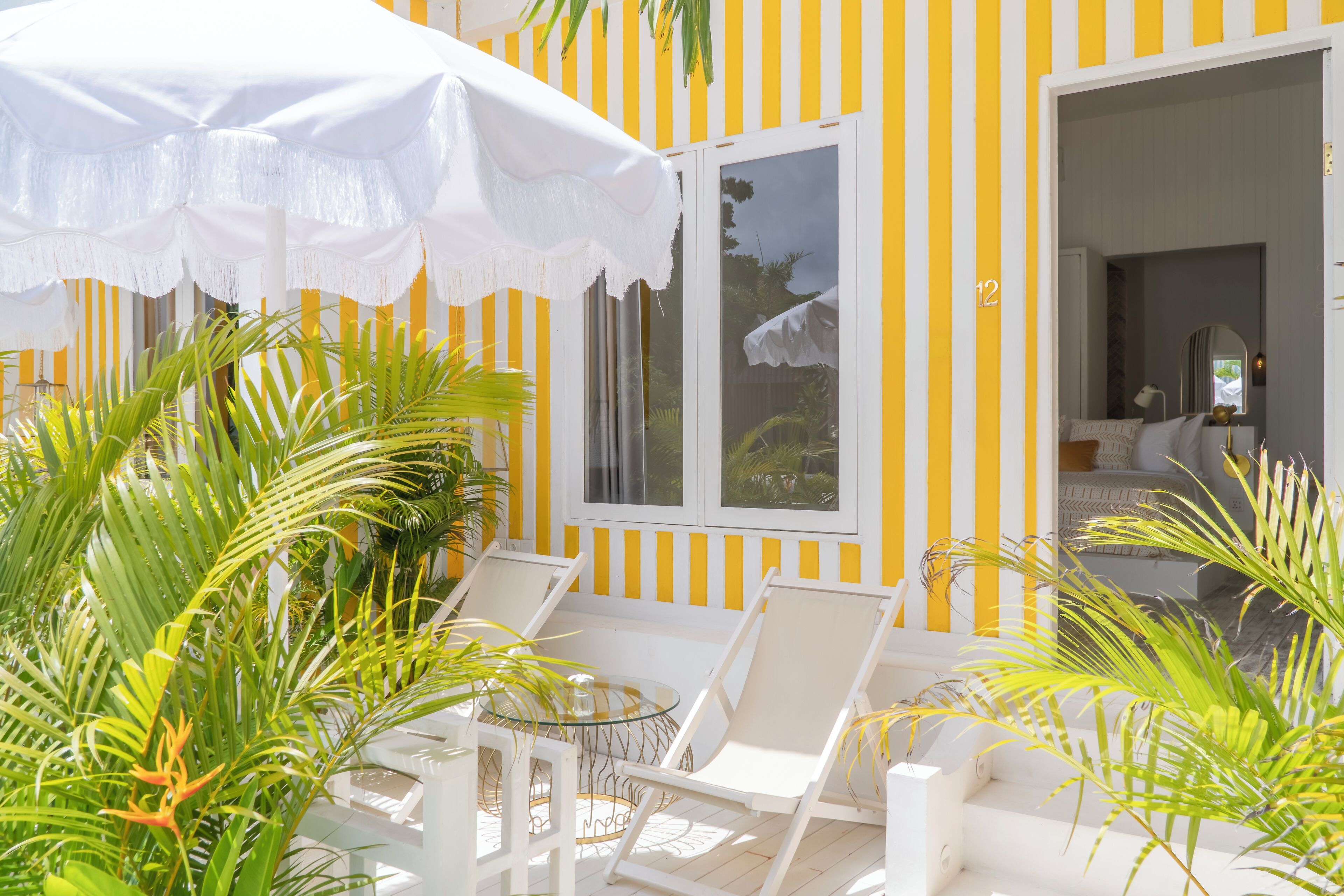 A sunlit outdoor seating area with white chairs and a white umbrella, surrounded by lush greenery.