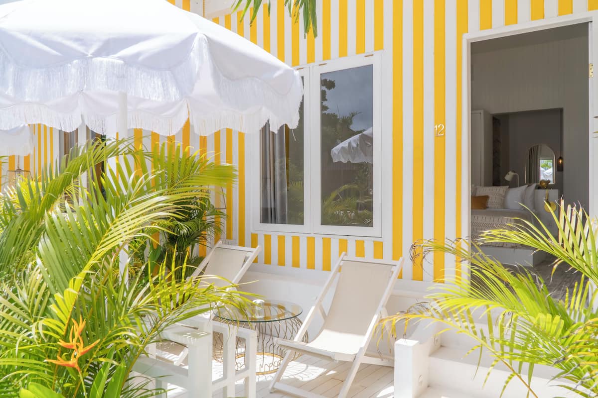 A sunlit outdoor seating area with white chairs and a white umbrella, surrounded by lush greenery.