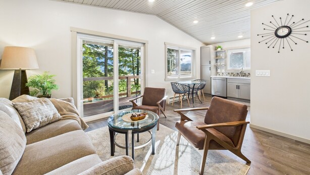 A spacious living area with a large sofa, a glass coffee table, and a dining table with chairs. There is a kitchen area with cabinets and a sink. The room has large windows with a view of trees and a deck. - Sunny Point 2BR with Deck & Mountain Views (Juneau)