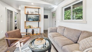 A modern living room with a beige sofa, a glass coffee table, and a brown leather chair. There is a bookshelf with decorative items and a painting on the wall. - Sunny Point 2BR with Deck & Mountain Views (Juneau)