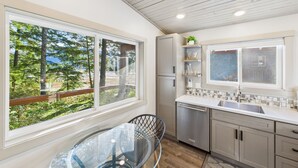 A modern kitchen with a view of a forest, a glass table, and a built-in dishwasher. - Sunny Point 2BR with Deck & Mountain Views (Juneau)