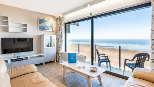 A modern living room with a TV, seating, and a table. A large window offers a view of the beach.