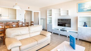 A modern living room with a beige sofa, a dining area with wooden chairs, a TV mounted on the wall, and a kitchen area with white cabinets.