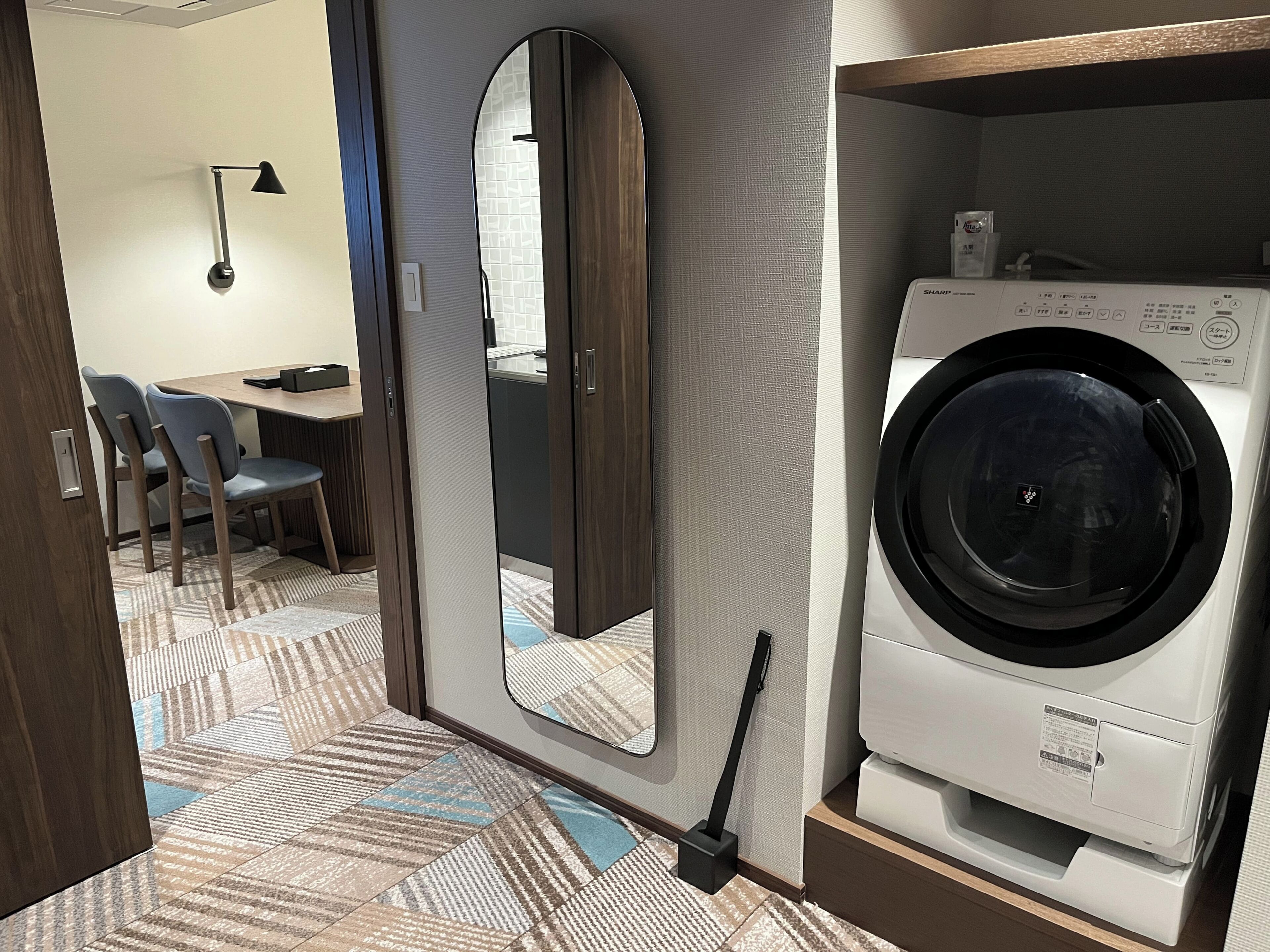 Hotelkamer met een wasmachine, een spiegel, een zithoek met stoelen en een vloer met een patroon.