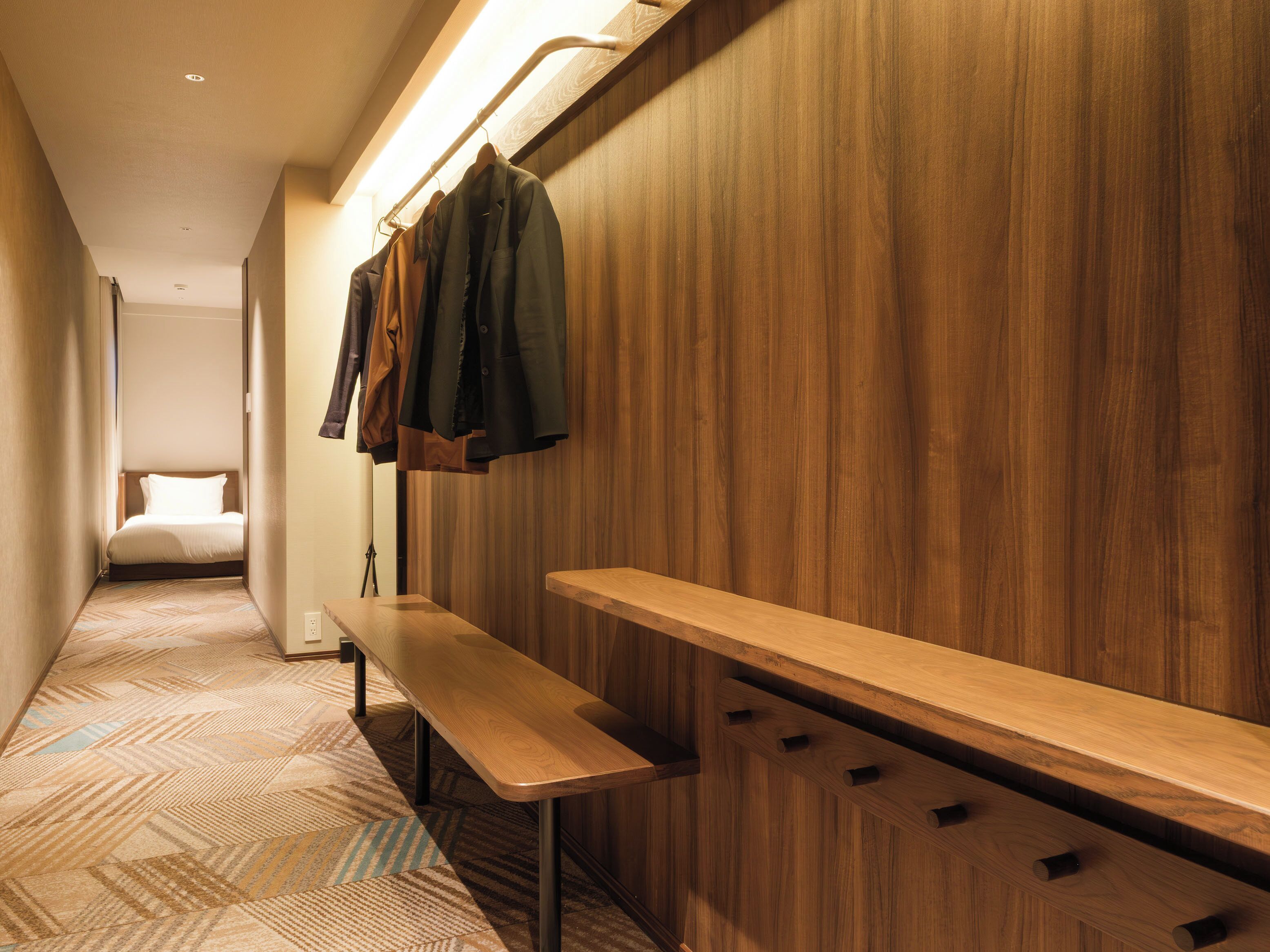 A hotel hallway with a bench, wooden wall paneling, and a bed in the distance.