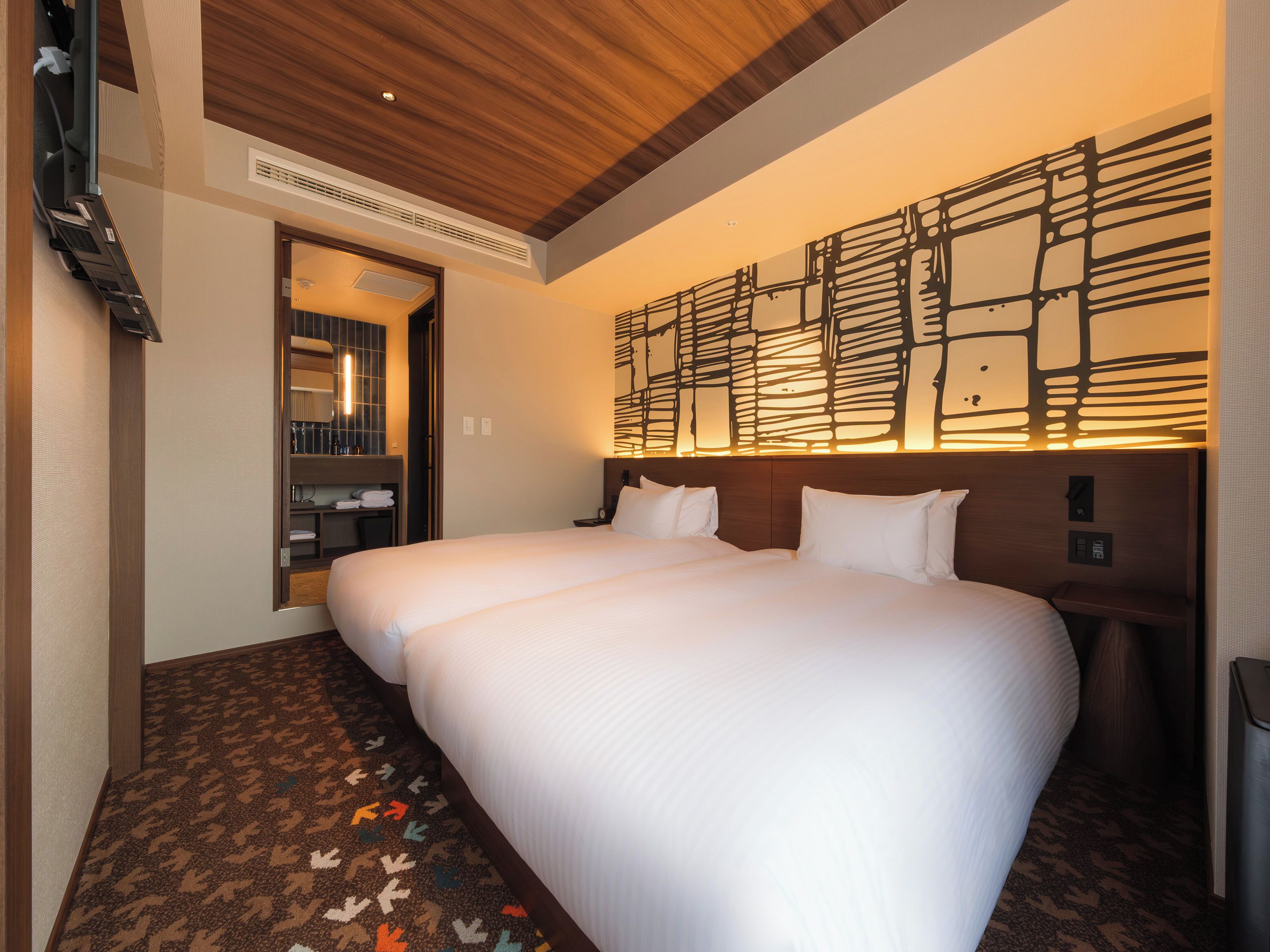 A hotel room with a large bed, a wooden ceiling, a wall-mounted light fixture, and a built-in shelf with towels.