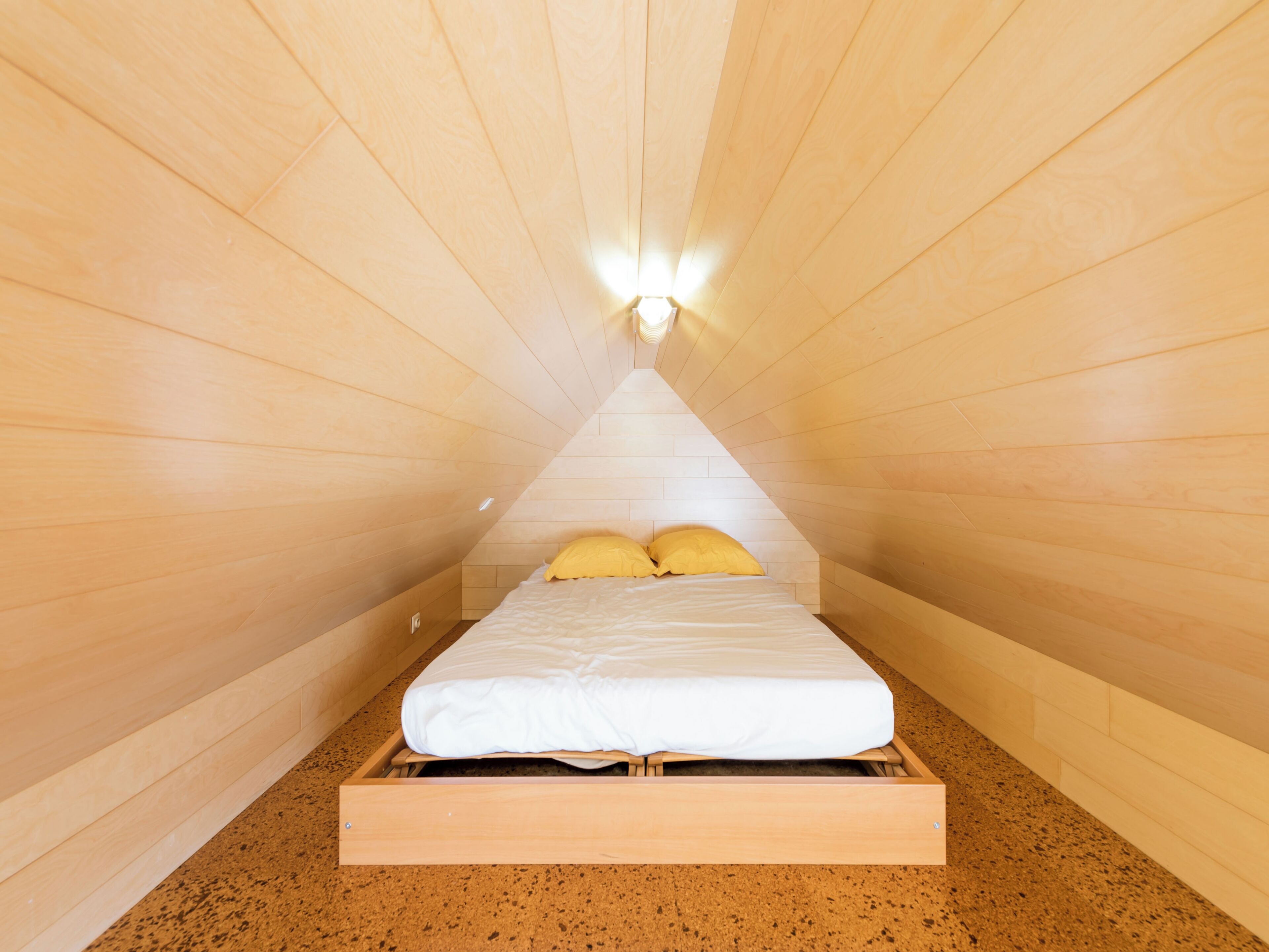 Une petite chambre mansardée de forme triangulaire, avec un plafond en pente en bois et un lit simple agrémenté d’oreillers jaunes.