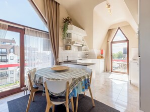 Zona pranzo con tavolo apparecchiato per due, cucina adiacente e vista all'esterno tramite ampie finestre.