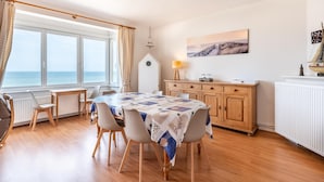 A dining area with a table and chairs, a view of the sea through large windows, a wooden cabinet, and a lamp. - Spacious family flat with sea view (Koksijde)
