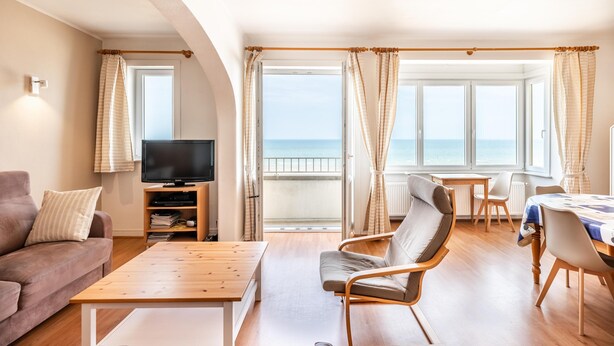A living room with a sofa, a TV, a wooden table, and a chair. There are large windows with curtains, a balcony, and a view of the sea. - Spacious family flat with sea view (Koksijde)