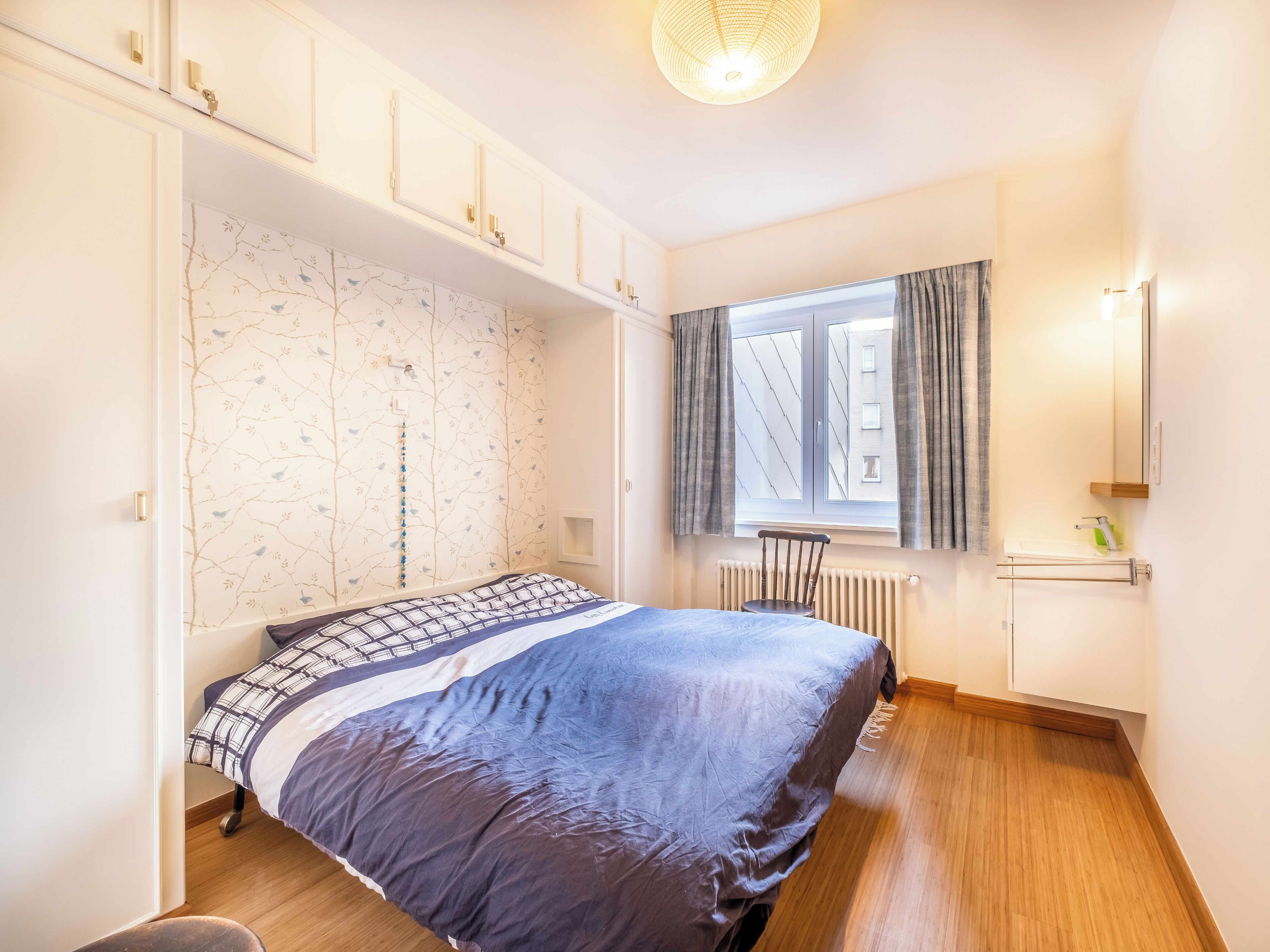 A bedroom with a bed, a window with curtains, a radiator, and a wardrobe.