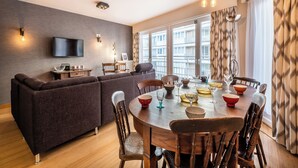 Een moderne woonkamer met een bank, eettafel en stoelen. Er is een televisie aan de muur bevestigd en een raam met gordijnen.