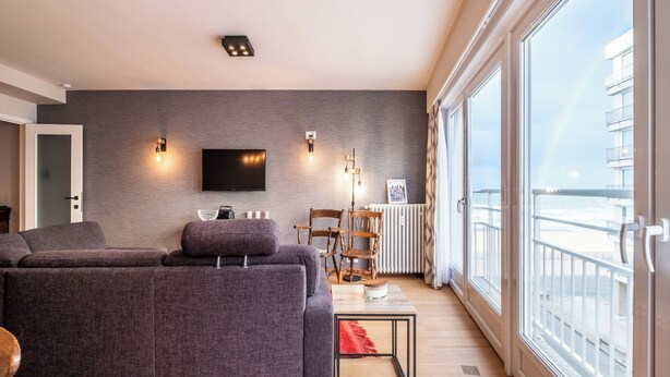 Een moderne woonkamer met een grijze bank, een aan de muur bevestigde televisie en uitzicht op zee door grote ramen.