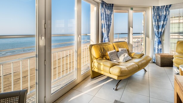 Ein geräumiger Balkon mit gelbem Sofa, großen Fenstern und Blick auf den Strand.