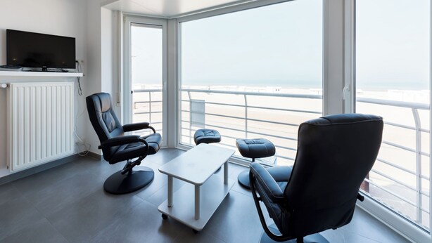 A modern living room with a large window, a flat-screen TV, a sofa, a coffee table, and a chair. - Stylish flat with sea view (Koksijde)