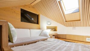 Una habitación acogedora con techo inclinado, una cama con colchón y almohadas blancas, una mesita de noche pequeña y una ventana.