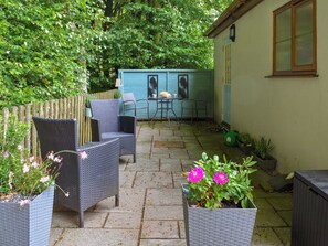 Een terras met rieten meubels, een tafel en stoelen. Er staan potplanten en een kleine schuur op de achtergrond.