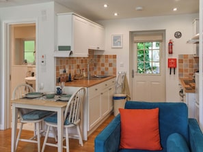 Une cuisine de taille réduite avec des meubles blancs, un plan de travail en bois et un évier. On y trouve un coin repas avec une table et des chaises, ainsi qu’un canapé bleu agrémenté d’un coussin orange. Une couverture anti-feu et un extincteur sont fixés au mur.