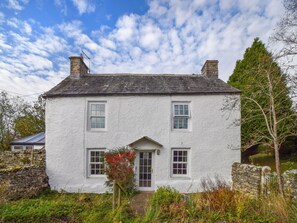 Une maison blanche à deux étages, avec des fondations en pierres apparentes, une entrée centrale et un petit jardin.