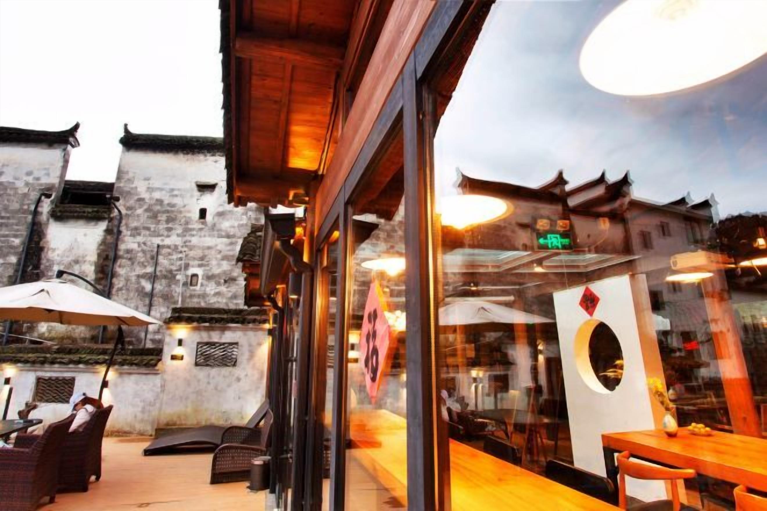 An outdoor seating area with tables and chairs, a glass wall reflecting the interior, and traditional architecture in the background.