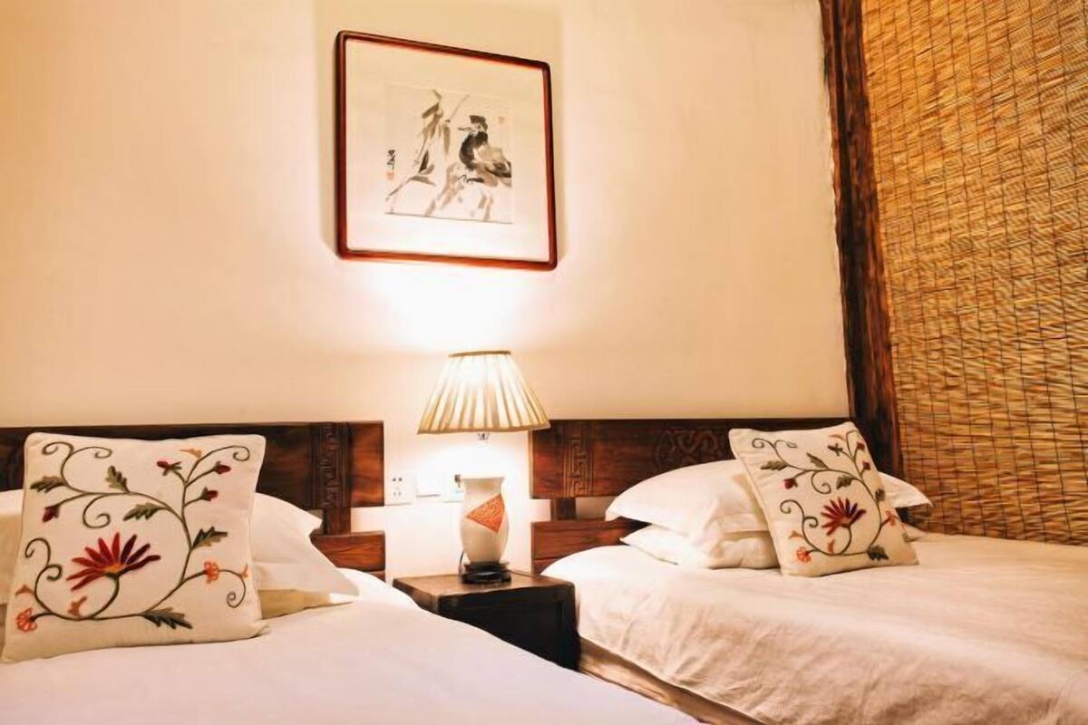 A hotel room with two beds, a nightstand with a lamp, a framed artwork on the wall, and a woven curtain.