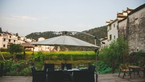 Ένας υπαίθριος χώρος με τραπέζι και καρέκλες κάτω από μια πέργκολα, με θέα ένα χωριό που διαθέτει παραδοσιακά κτίρια και ένα καταπράσινο τοπίο.