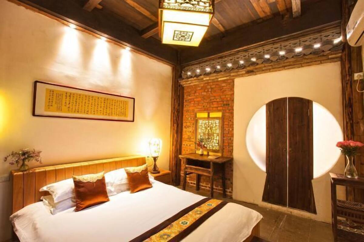 A hotel room with a wooden bed, a desk, a wall-mounted lamp, and a large circular mirror.